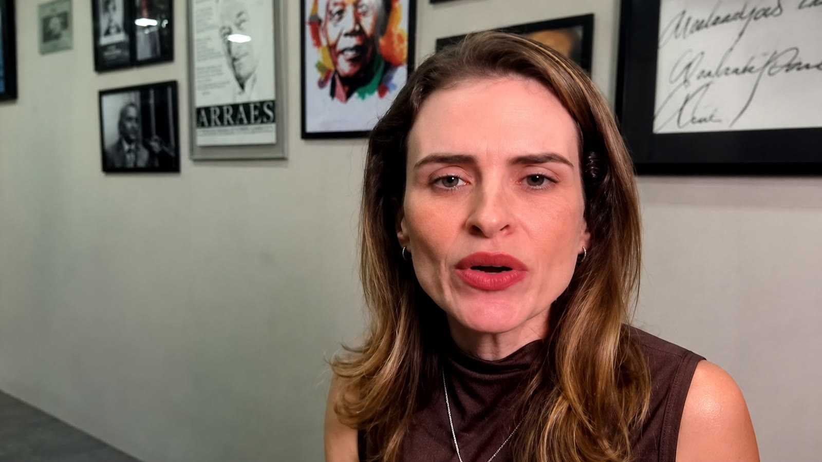 A candidata ao Senado Marilia Arraes durante entrevista, diante de uma parede com retratos e um cartaz de Miguel Arraes